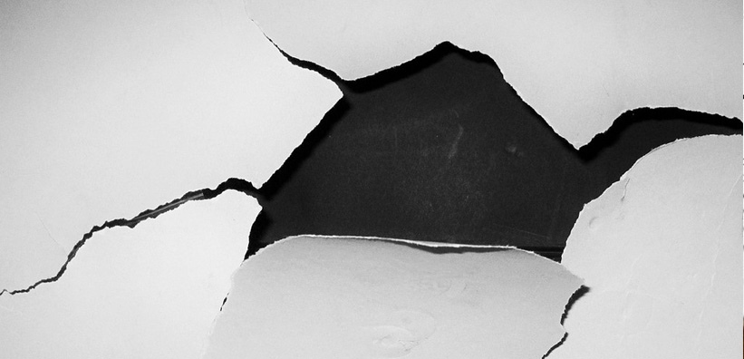 Das Bild zeigt eine Oberfläche, die rissig ist und abgeplatzte Bereiche aufweist, wodurch ein dunklerer Untergrund sichtbar wird.