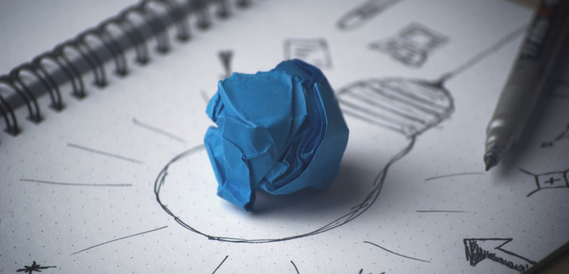 Das Bild zeigt einen zerknüllten blauen Papierball, der auf einem Notizbuch liegt, das eine gezeichnete Glühbirne und andere Skizzen enthält.