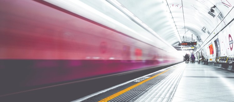 Das Bild zeigt einen Zug, der mit hoher Geschwindigkeit durch einen U-Bahn- oder Bahntunnel fährt. Die Bewegungsunschärfe des Zuges deutet auf eine Langzeitbelichtung hin, während die Plattform und der Tunnel scharf abgebildet sind.