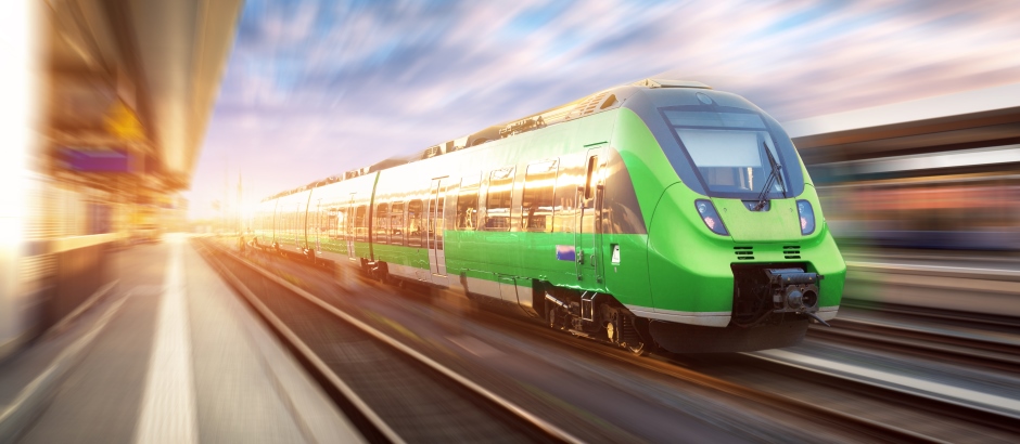 Ein moderner, grüner Zug fährt mit hoher Geschwindigkeit durch einen Bahnhof. Die Bewegung ist deutlich sichtbar durch verschwommene Linien im Hintergrund. Die Sonne scheint von links ins Bild und erzeugt eine warme, dynamische Atmosphäre – die Szene symbolisiert Fortschritt und Mobilität.