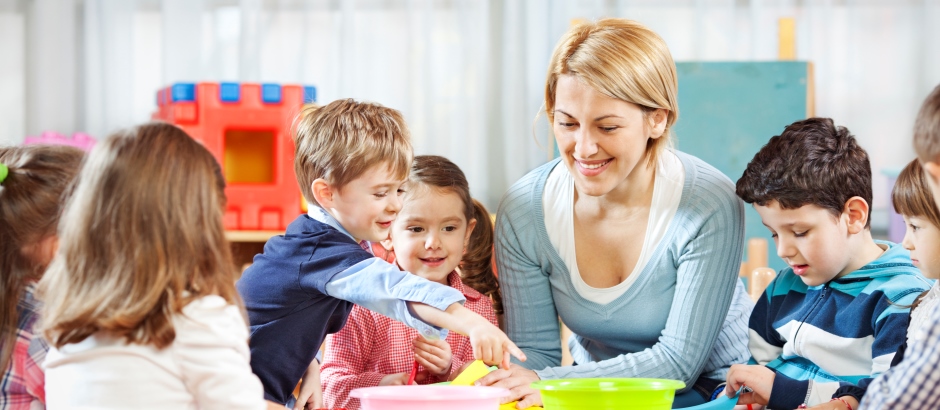 Eine Erzieherin sitzt mit mehreren kleinen Kindern an einem Tisch. Alle schauen fröhlich und spielen gemeinsam mit buntem Spielmaterial. Die Stimmung ist freundlich und lebendig – eine typische Szene aus dem Kindergartenalltag.