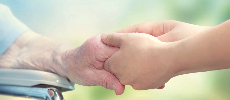 Eine ältere Frau im Rollstuhl hält die Hand einer jungen Betreuerin.