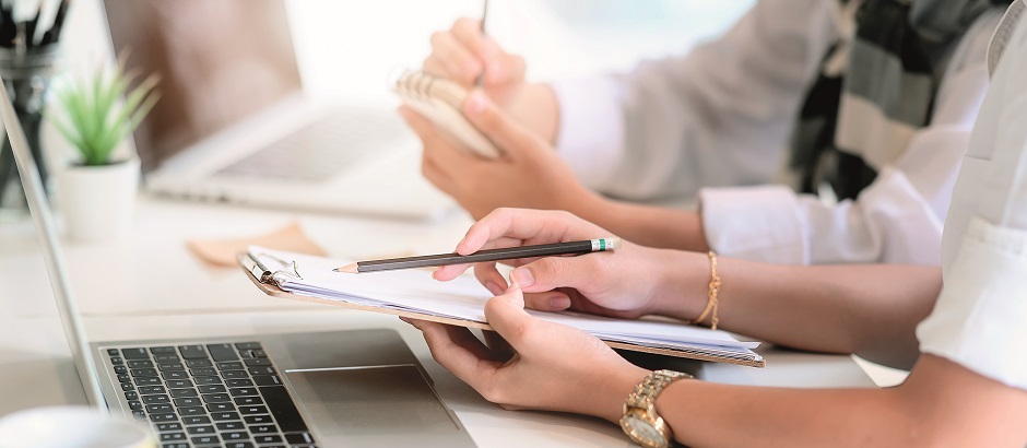 Zwei Personen schreiben sich Notizen auf. Am Schreibtisch steht ein Laptop.