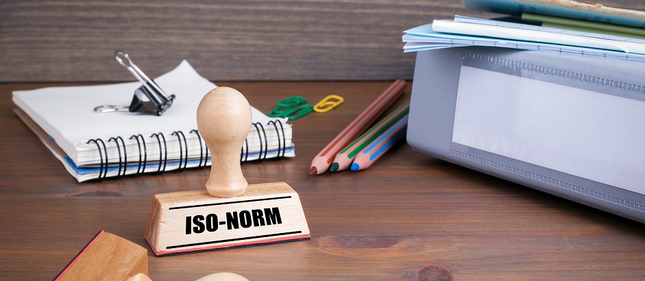 Ein Schreibtisch, auf welchem Aktenordner, Buntstifte, Notizblöcke, Klammern und ein ISO-Norm-Stempel liegen